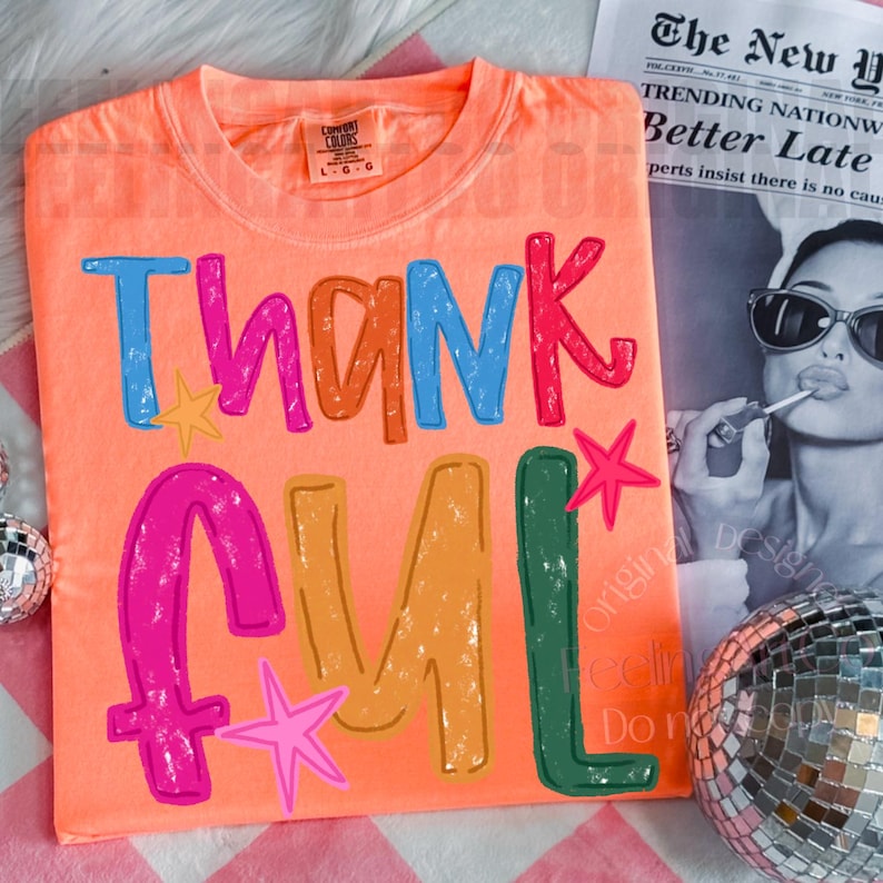 May include: A bright orange crew neck t-shirt featuring the text Thankful in large, multi-colored, distressed-style letters. The design includes small pink and yellow star graphics interspersed among the colorful, bold, hand-drawn style typography.