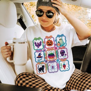 May include: White t-shirt featuring a colorful Halloween-themed graphic. The design includes various spooky icons like a bat, pumpkin, ghost, witch hat, cauldron, skull, black cat, spider, and fish. The person is wearing a gray beanie, sunglasses, and a gold chain.