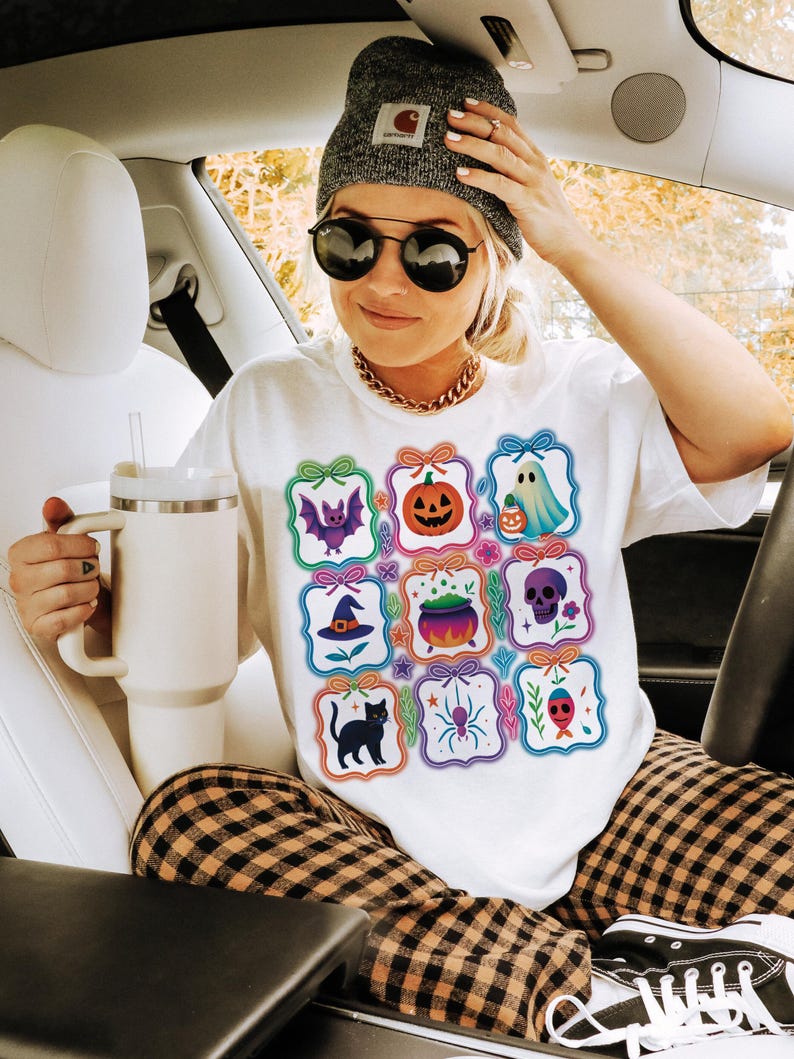May include: White t-shirt featuring a colorful Halloween-themed graphic. The design includes various spooky icons like a bat, pumpkin, ghost, witch hat, cauldron, skull, black cat, spider, and fish. The person is wearing a gray beanie, sunglasses, and a gold chain.