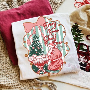 Puede incluir: Camiseta blanca con un gr&aacute;fico navide&ntilde;o. El dise&ntilde;o presenta un &aacute;rbol de Navidad, Pap&aacute; Noel arrodillado y el texto "Every Knee Shall Bow" en un marco decorativo. Una camiseta roja est&aacute; doblada al lado.
