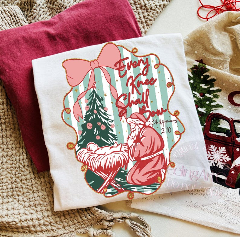 Puede incluir: Camiseta blanca con un gr&aacute;fico navide&ntilde;o. El dise&ntilde;o presenta un &aacute;rbol de Navidad, Pap&aacute; Noel arrodillado y el texto "Every Knee Shall Bow" en un marco decorativo. Una camiseta roja est&aacute; doblada al lado.