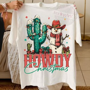 Peut inclure: T-shirt blanc avec un motif de No&euml;l fantaisiste. Le graphisme comprend un cactus d&eacute;cor&eacute; de guirlandes lumineuses, un bonhomme de neige portant un chapeau de cowboy et une &eacute;charpe, et les mots "HOWDY Christmas" dans une police festive.