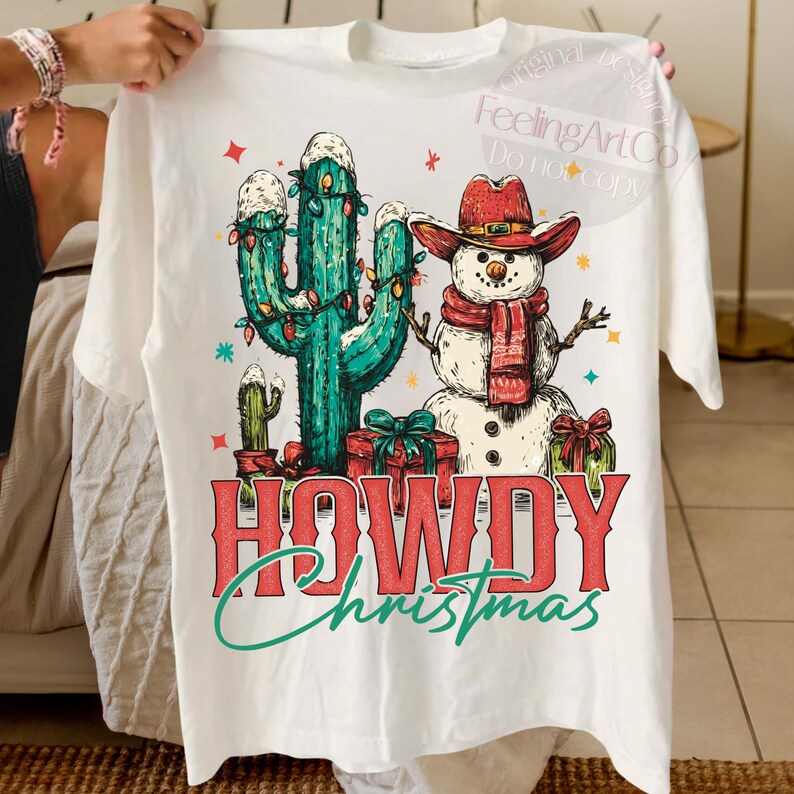 Peut inclure: T-shirt blanc avec un motif de No&euml;l fantaisiste. Le graphisme comprend un cactus d&eacute;cor&eacute; de guirlandes lumineuses, un bonhomme de neige portant un chapeau de cowboy et une &eacute;charpe, et les mots "HOWDY Christmas" dans une police festive.