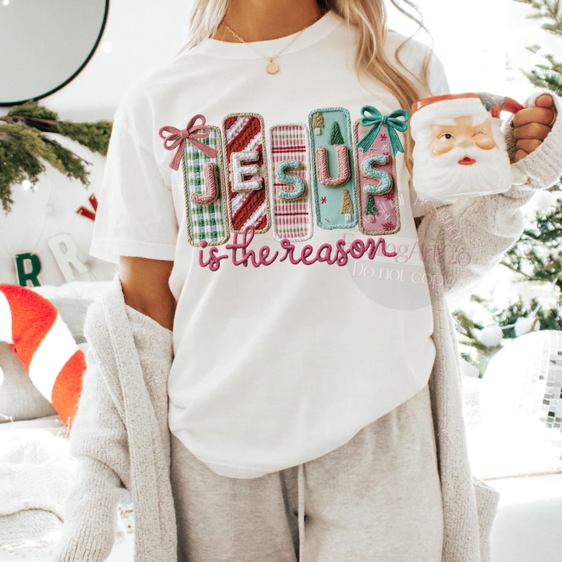 May include: White t-shirt with the text "JESUS is the reason" in a colorful, festive design. The design features the word "JESUS" in a stacked format, with each letter decorated with Christmas-themed patterns and bows. A Santa mug is held in the hand.