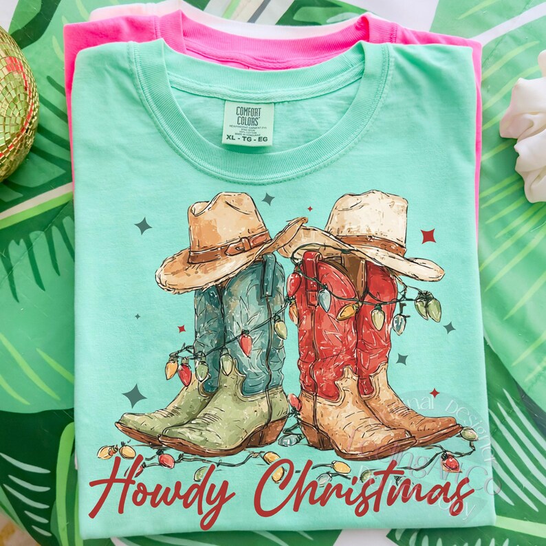 May include: A light green t-shirt with a graphic of cowboy boots and hats decorated with Christmas lights. The text "Howdy Christmas" is printed in red. The shirt is folded on top of a pink shirt.
