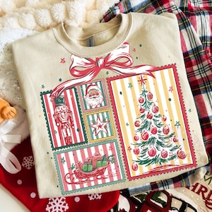 May include: A beige Christmas-themed sweatshirt featuring a large red bow and several postage stamp-style images. The images include a nutcracker, Santa Claus, a star, a Christmas tree, and a sleigh with gifts. The design has red, green, and yellow accents.