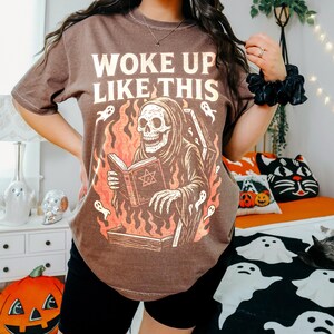 May include: Brown t-shirt featuring the text "WOKE UP LIKE THIS" and a graphic of a skeleton in a robe reading a book, with flames and ghosts. The shirt is worn with black shorts.