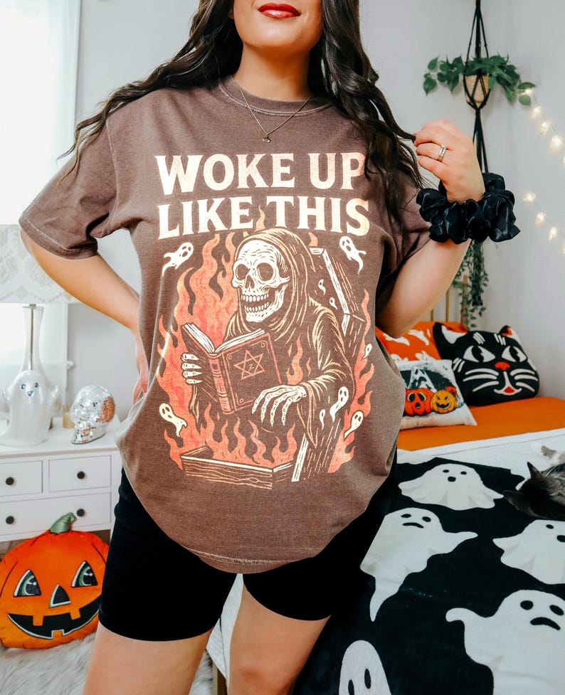 May include: Brown t-shirt featuring the text "WOKE UP LIKE THIS" and a graphic of a skeleton in a robe reading a book, with flames and ghosts. The shirt is worn with black shorts.