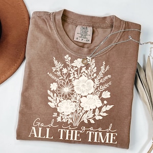 Könnte beinhalten: Ein braunes T-Shirt mit einem weißen Blumenmuster und dem Text "God is good ALL THE TIME".