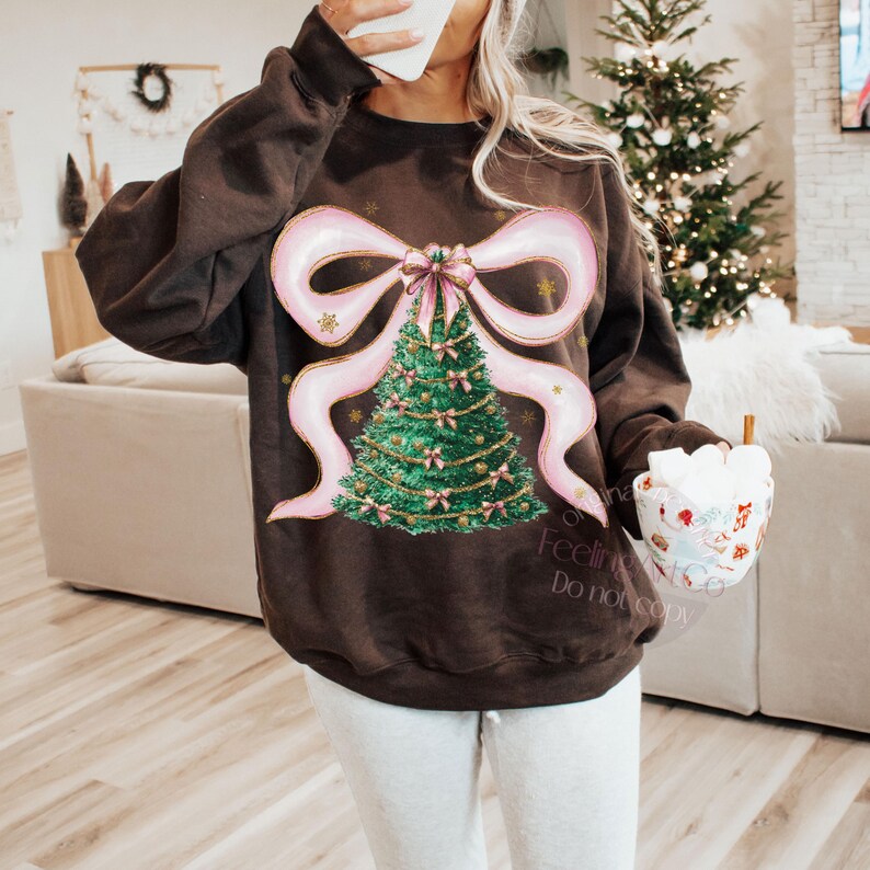 Pu&ograve; includere: Una felpa marrone con un design a forma di albero di Natale. Il design include un fiocco rosa, un albero di Natale verde e dettagli dorati. Il testo "Feeling Cozy" &egrave; visibile in basso a destra.