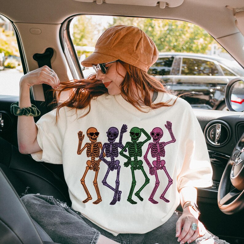 Puede incluir: Sudadera de color crema con un gr&aacute;fico colorido de cinco esqueletos bailando. Cada esqueleto es de un color diferente: naranja, morado, verde y rosa. Los esqueletos llevan gafas de sol y saludan. La sudadera la lleva una persona en un coche.