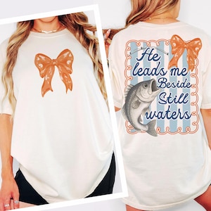 May include: Cream-colored t-shirt featuring an orange bow on the front. The back of the shirt displays a blue and white striped design with the text "He leads me Beside Still waters" and a fish illustration.