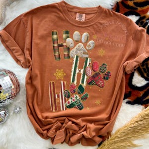 K&ouml;nnte beinhalten: Gebrannt orangefarbenes T-Shirt mit dem Text "Ho Ho Ho" in einem Patchwork-Design mit Pfotenabdr&uuml;cken und Schneeflocken. Die Buchstaben sind in gr&uuml;nen, roten und wei&szlig;en Karomustern gehalten. Das Shirt hat einen Rundhalsausschnitt und kurze &Auml;rmel.