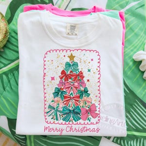 May include: White t-shirt featuring a Christmas tree design made of bows in red, pink, and teal. The design includes gold accents and a gold star on top. The words "Merry Christmas" are written in pink script at the bottom. The shirt is folded with pink and green shirts underneath.