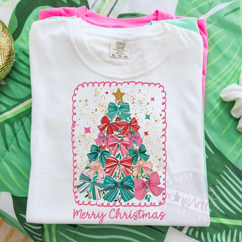 May include: White t-shirt featuring a Christmas tree design made of bows in red, pink, and teal. The design includes gold accents and a gold star on top. The words "Merry Christmas" are written in pink script at the bottom. The shirt is folded with pink and green shirts underneath.