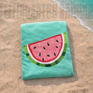 May include: A turquoise t-shirt with a watermelon slice applique. The watermelon features a red and white gingham pattern, white trim, green rind, and black seed details. The shirt is lying on a sandy beach.