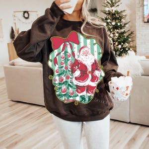 May include: A dark brown sweatshirt featuring a Christmas-themed graphic. The graphic includes a red bow, a Christmas tree with ornaments, and Santa Claus in a red suit. The sweatshirt is a cozy, festive apparel item.