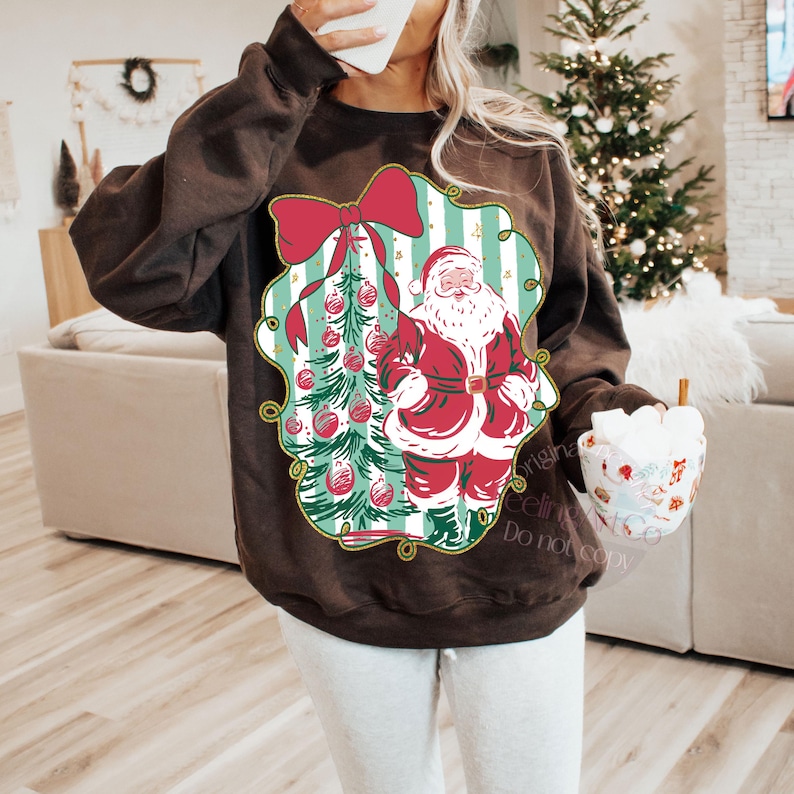 May include: A dark brown sweatshirt featuring a Christmas-themed graphic. The graphic includes a red bow, a Christmas tree with ornaments, and Santa Claus in a red suit. The sweatshirt is a cozy, festive apparel item.