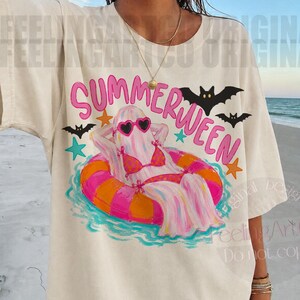 K&ouml;nnte beinhalten: Cremefarbenes T-Shirt mit einem "Summerween"-Motiv. Das Design zeigt einen Geist mit Sonnenbrille und Bikini, der in einem rosa-orangenen Schwimmring entspannt. Schwarze Flederm&auml;use und Sterne umgeben den Geist.