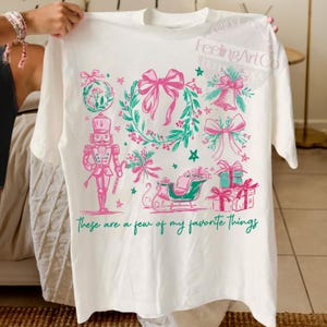 May include: White t-shirt with a festive design featuring pink bows, a wreath, a nutcracker, and a sleigh. The text "These are a few of my favorite things" is printed in green. The design includes stars and bell accents.