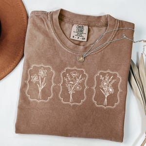 May include: A brown t-shirt with a white graphic of three floral bouquets in frames. The text "COMFORT COLORS" is visible on the shirt.