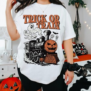 May include: White t-shirt with a Halloween graphic. The design shows a train with a pumpkin face, ghosts, and the text "Trick or Train" in orange. A fun and festive tee for the spooky season.