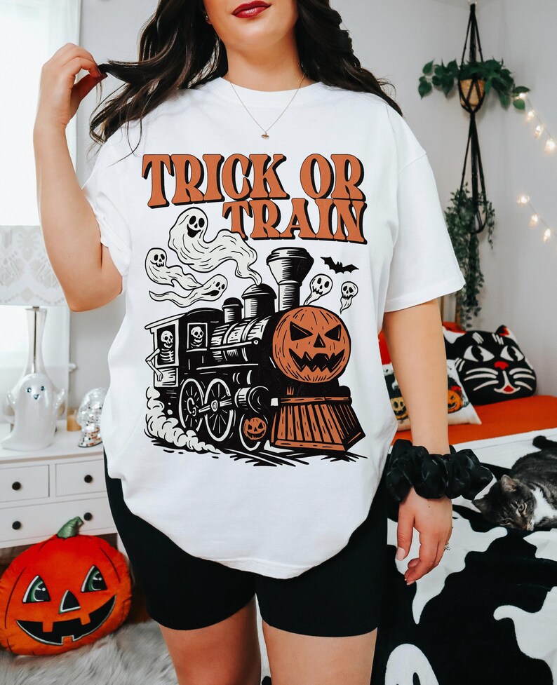 May include: White t-shirt with a Halloween graphic. The design shows a train with a pumpkin face, ghosts, and the text "Trick or Train" in orange. A fun and festive tee for the spooky season.