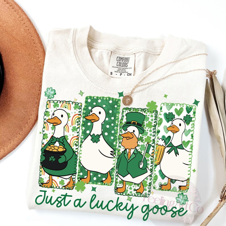 May include: Cream-colored t-shirt with a St. Patrick's Day theme. Features four cartoon geese in green frames, each with a different festive accessory. The text "Just a lucky goose" is printed in green script.