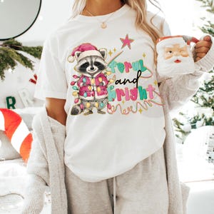 May include: White t-shirt featuring a cartoon raccoon wearing a Santa hat and a pink jacket adorned with Christmas lights. The text "Feral and Bright" is written in colorful letters, with a star and decorative lines.
