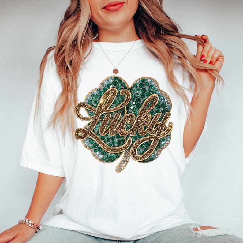 May include: White t-shirt featuring a large, embellished shamrock design. The shamrock is green with gold accents and the word "Lucky" in gold script. The shirt is a casual style.