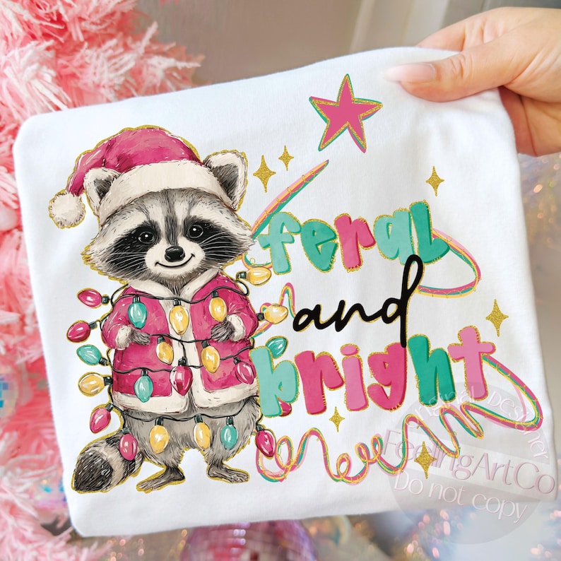 May include: White t-shirt featuring a cartoon raccoon wearing a Santa hat and a pink jacket adorned with Christmas lights. The text "feral and bright" is written in colorful letters with a star and sparkles.