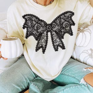 May include: Cream-colored t-shirt featuring a large black lace bow design. The bow has intricate detailing and a scalloped edge. The shirt is paired with light blue jeans. A gold necklace is visible.
