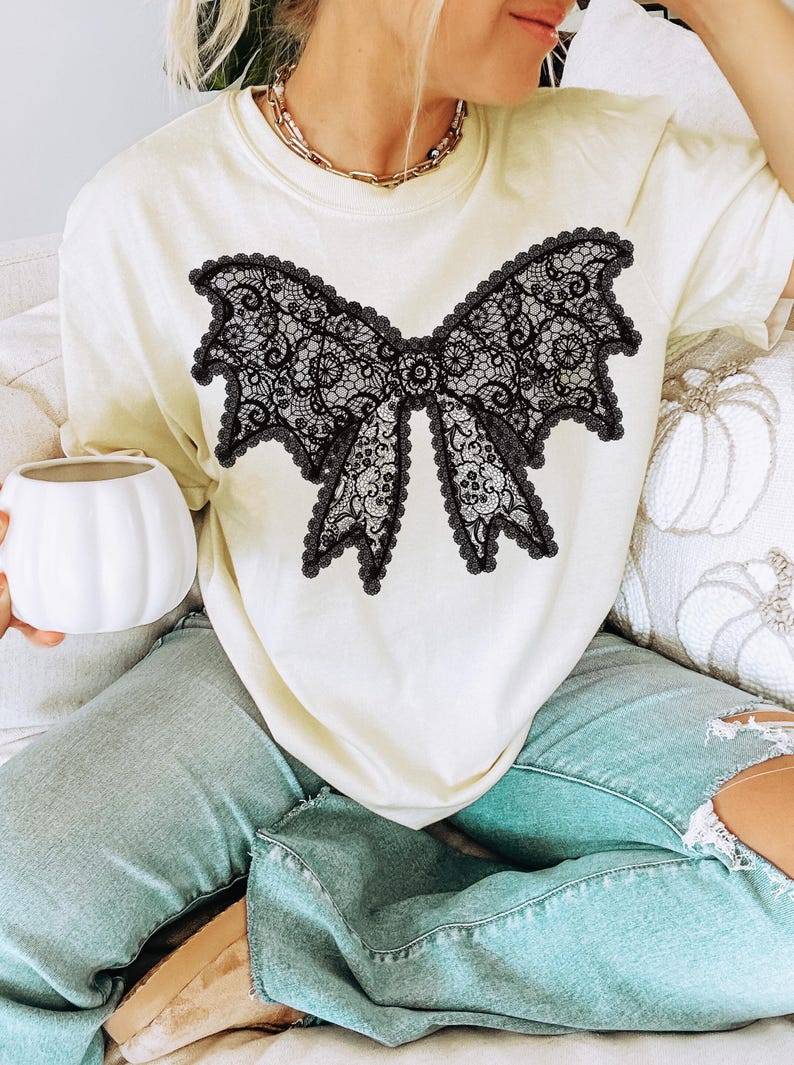 May include: Cream-colored t-shirt featuring a large black lace bow design. The bow has intricate detailing and a scalloped edge. The shirt is paired with light blue jeans. A gold necklace is visible.