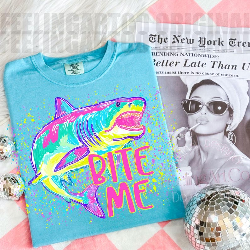 Op de afbeelding: Een lichtblauw t-shirt met een kleurrijke haai-afbeelding en de tekst "BITE ME" in roze. De haai is afgebeeld in tinten roze, groen, geel en blauw. Het shirt heeft een ronde hals en korte mouwen.