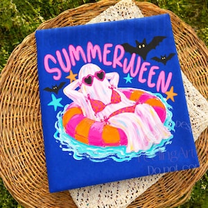 K&ouml;nnte beinhalten: Blaues T-Shirt mit dem Wort "SUMMERWEEN" in Pink und Wei&szlig;. Die Grafik zeigt einen Geist mit herzf&ouml;rmiger Sonnenbrille und Bikini, der auf einem rosa-orangenen Schwimmring mit Flederm&auml;usen und Sternen schwimmt.