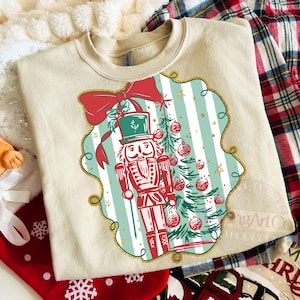 May include: Cream-coloured sweatshirt with a festive design. The graphic features a red and green nutcracker soldier, a Christmas tree, and a red bow. The design is set against a striped background with gold accents, ideal for the holiday season.