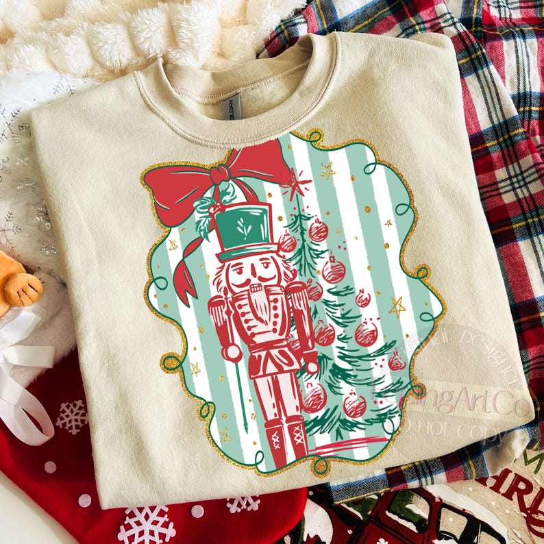 May include: Cream-coloured sweatshirt with a festive design. The graphic features a red and green nutcracker soldier, a Christmas tree, and a red bow. The design is set against a striped background with gold accents, ideal for the holiday season.