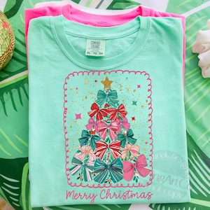May include: A light green t-shirt featuring a Christmas tree design made of bows in pink, red, and teal. The words "Merry Christmas" are printed in pink. Another pink t-shirt is stacked underneath.