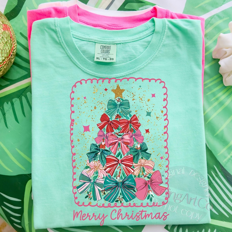 May include: A light green t-shirt featuring a Christmas tree design made of bows in pink, red, and teal. The words "Merry Christmas" are printed in pink. Another pink t-shirt is stacked underneath.