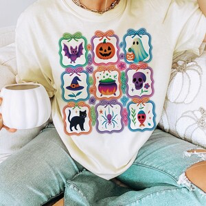 May include: Cream-colored t-shirt featuring a grid of Halloween-themed illustrations. Each square showcases a different design, including a bat, pumpkin, ghost, witch's hat, cauldron, skull, black cat, spider, and a fish-like creature.