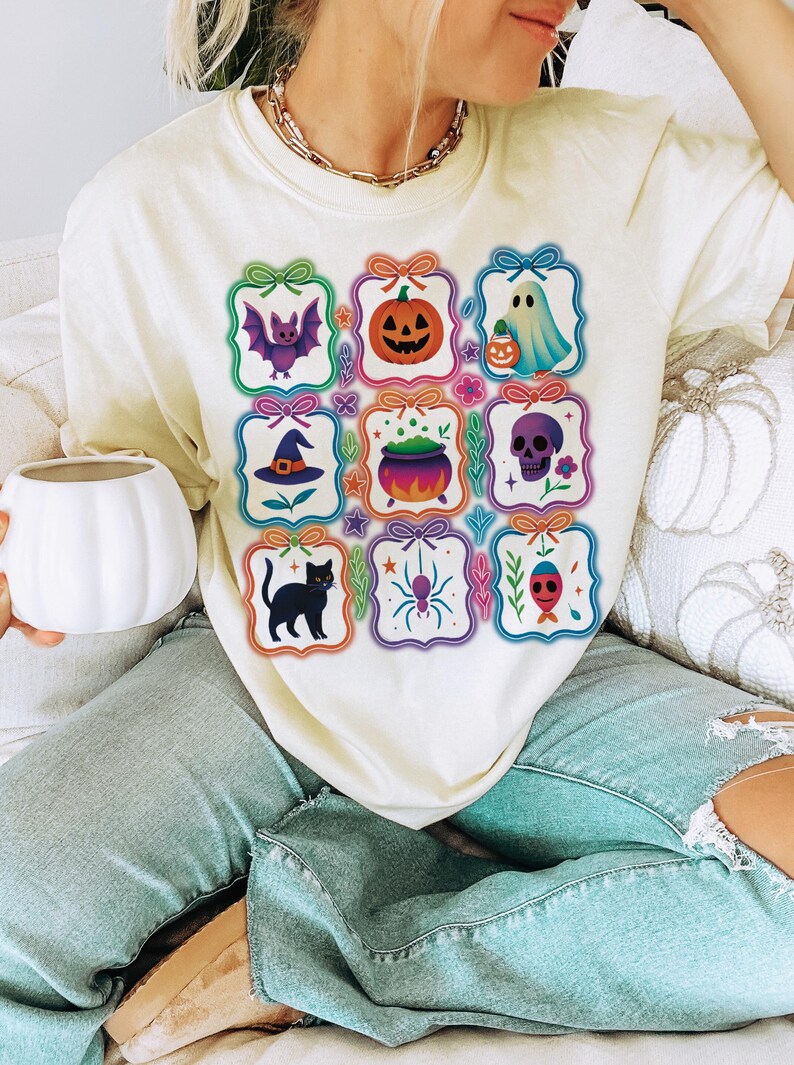 May include: Cream-colored t-shirt featuring a grid of Halloween-themed illustrations. Each square showcases a different design, including a bat, pumpkin, ghost, witch's hat, cauldron, skull, black cat, spider, and a fish-like creature.