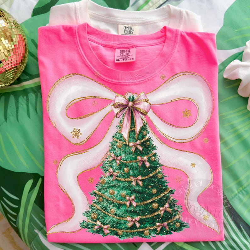 Pu&ograve; includere: Una maglietta rosa con un disegno di albero di Natale, un grande fiocco bianco e dettagli dorati. L'albero &egrave; verde con fiocchi rosa e ornamenti dorati. Una maglietta bianca &egrave; visibile dietro la maglietta rosa.