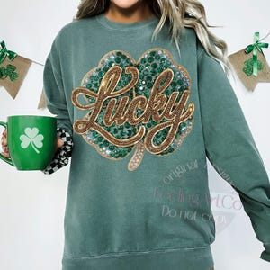 May include: Green sweatshirt with a large, sparkling shamrock design. The word "Lucky" is written in gold script across the shamrock. A green mug with a shamrock is held in the image. The sweatshirt has a relaxed fit.
