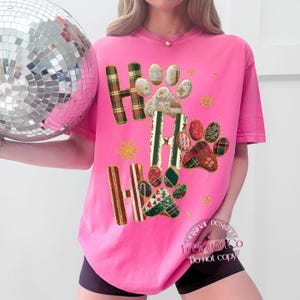K&ouml;nnte beinhalten: Ein leuchtend pinkfarbenes T-Shirt mit den Worten "HO HO HO" in einem Karomuster und Pfotenabdruck-Design. Das Shirt hat eine lockere Passform und kurze &Auml;rmel. Goldene Glitzer-Schneeflocken sind um das Design herum verstreut.