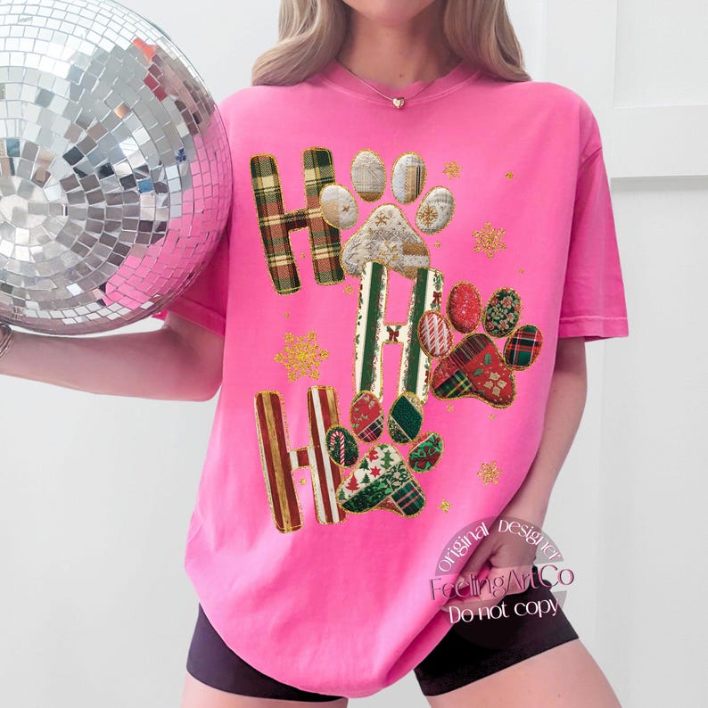 K&ouml;nnte beinhalten: Ein leuchtend pinkfarbenes T-Shirt mit den Worten "HO HO HO" in einem Karomuster und Pfotenabdruck-Design. Das Shirt hat eine lockere Passform und kurze &Auml;rmel. Goldene Glitzer-Schneeflocken sind um das Design herum verstreut.