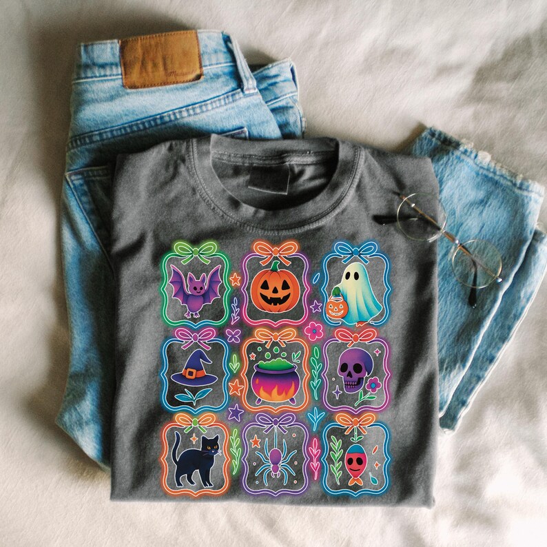 May include: Gray t-shirt featuring Halloween-themed graphics. The shirt displays nine colorful illustrations of a bat, pumpkin, ghost, witch hat, cauldron, skull, black cat, spider, and fish, each within a neon-outlined frame. The shirt is paired with blue jeans.