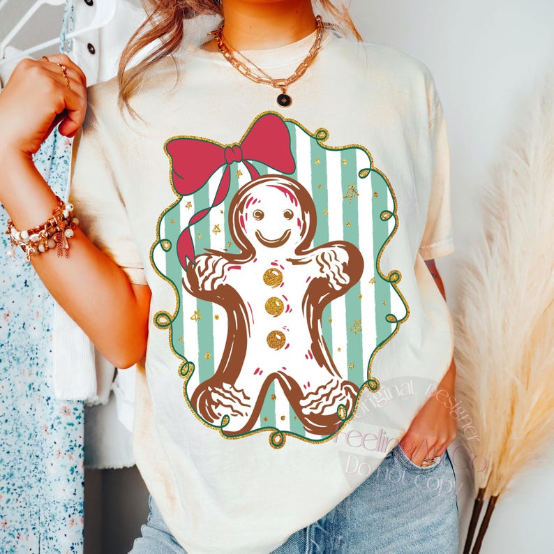 May include: Cream-colored t-shirt featuring a gingerbread man design. The gingerbread man is outlined in brown with white frosting and gold accents. A red bow sits above the gingerbread man, and the design is set against a teal and white striped background.