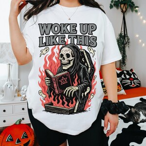 May include: White t-shirt featuring the text "WOKE UP LIKE THIS" above a graphic of a skeleton in a grim reaper robe reading a book in a coffin surrounded by flames and ghosts. The shirt is worn with black shorts.