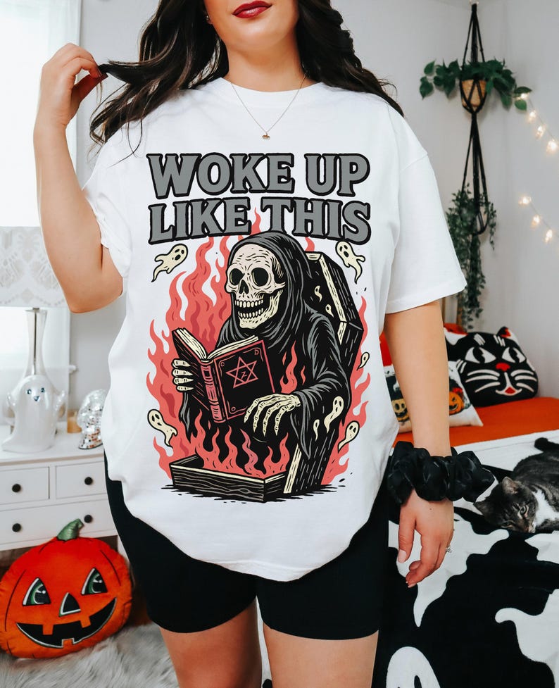 May include: White t-shirt featuring the text "WOKE UP LIKE THIS" above a graphic of a skeleton in a grim reaper robe reading a book in a coffin surrounded by flames and ghosts. The shirt is worn with black shorts.