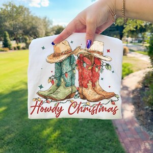 May include: White t-shirt featuring a watercolor illustration of cowboy boots adorned with Christmas lights and cowboy hats. The text "Howdy Christmas" is written in red script below the boots.